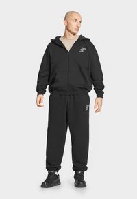 Czarna bluza z kapturem na zamek z troczkami, ściągaczami przy rękawach i kieszeniami z przodu, zestawiona z pasującymi spodniami dresowymi z logo na udzie.