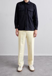 Navy button-up shirt with two chest pockets, paired with pale yellow pleated trousers and white sneakers with brown soles.