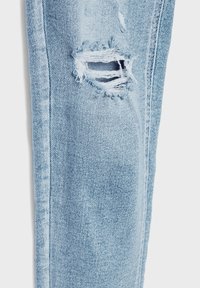 Jambe de jean en denim bleu clair avec une déchirure horizontale montrant des fils blancs effilochés et des bords de tissu.