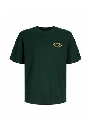 Ung person med krøllet hår iført en mørkegrøn Jack & Jones t-shirt, brune bukser og beige sko, stående med ryggen til på en ensfarvet baggrund.
