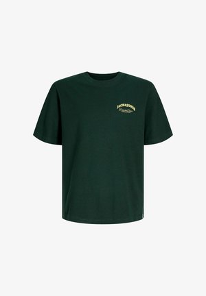 Ung person med krøllet hår iført en mørkegrøn Jack & Jones t-shirt, brune bukser og beige sko, stående med ryggen til på en ensfarvet baggrund.