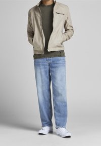 Béžová lehká bunda se zipovou kapsou na hrudi, pod ní olivově zelená košile s dlouhým rukávem, v kombinaci se světle modrými džíny rovného střihu a bílými botami.