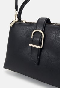 Sac à main en cuir noir avec fermeture éclair dorée, forme rectangulaire, et une bandoulière avant ornée d'une boucle dorée près du haut.