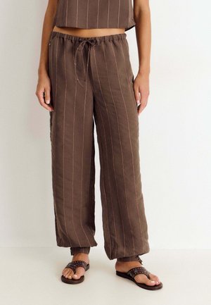 Pantalones marrones de pierna ancha con rayas verticales finas, cintura elástica con cordón, usados con sandalias marrón oscuro con tachuelas.