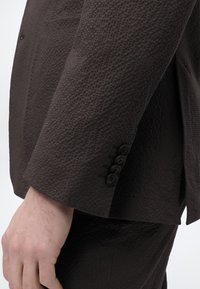 Κοντινό πλάνο από το χέρι ενός ατόμου που φοράει σκούρο υφασμάτινο σακάκι με τέσσερα μαύρα κουμπιά στη μανσέτα, πάνω σε απλό φόντο.