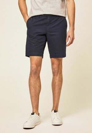 Hombre con pantalones cortos azul marino hasta la rodilla, camiseta gris claro y zapatillas blancas, de pie con las manos parcialmente en los bolsillos.