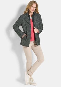 Vrouw in een gestructureerde donkergrijze jas, koraalkleurige trui, beige broek en witte sneakers, staand met één hand in haar zak en één been gekruist.