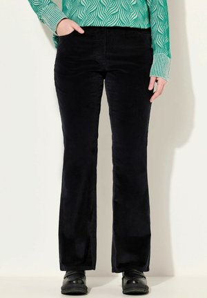 Kobieta ubrana w czarne spodnie sztruksowe i czarne buty, z ręką w kieszeni, w zielono-białą bluzkę z długim rękawem o wzorze.