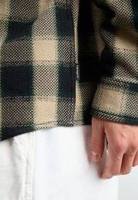Camisa de forro polar a cuadros beige y verde con detalles en negro, tela texturizada, una etiqueta lateral y mangas con puños, combinada con ropa blanca.