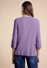 Top viola leggero con maniche tre quarti, vestibilità rilassata, scollatura arrotondata e orlo leggermente curvo. Indossato con jeans blu.