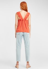 Rostorange ärmlös topp med volangärmar, öppen ryggdesign; kombinerad med ljusblå, något slitna, löst sittande jeans och beige klackar.