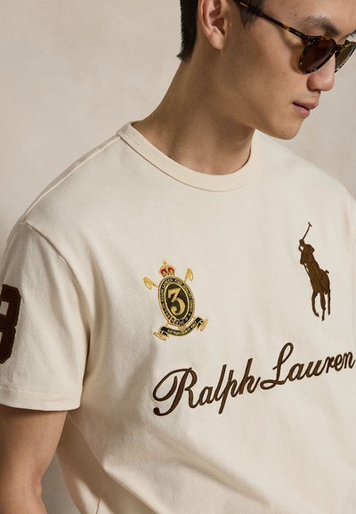 Άνδρας που φοράει μπεζ μπλουζάκι Ralph Lauren με μεγάλο λογότυπο, κεντημένο έμβλημα με τον αριθμό 3 και σκοτεινά γυαλιά ηλίου, στραμμένος στο πλάι.