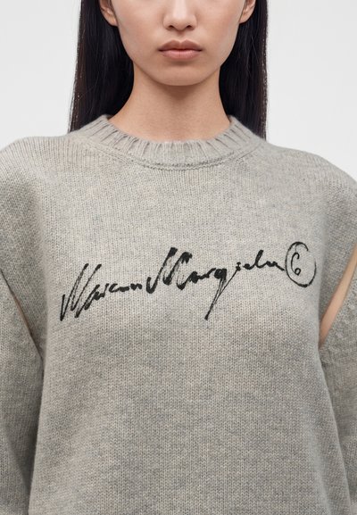 Jeune personne aux longs cheveux foncés portant un pull en maille gris avec un texte noir en italique sur la poitrine sur un fond uni.