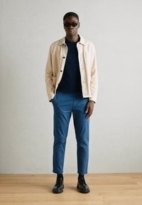 Világos színű dzseki, sötétkék pulóver, kék nadrág és fekete cipő. Kiegészítők között sötét napszemüveg található. Simák, laza dizájn.