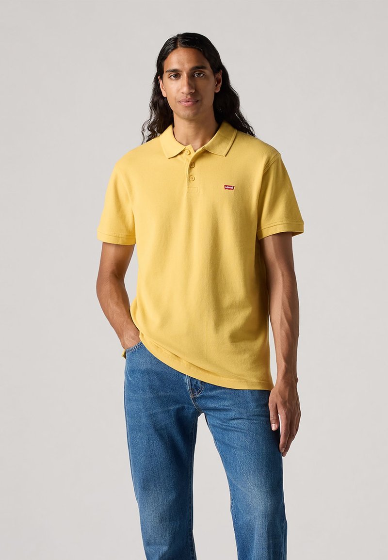 Polo shirt din bumbac galben, cu guler și mâneci scurte, având un mic logo roșu pe piept. Purtați cu blugi albaștri.