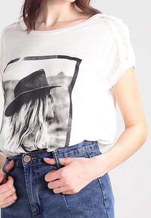 Camiseta blanca con una impresión gráfica en blanco y negro de una mujer con un sombrero, con detalles de encaje en los hombros, combinada con unos vaqueros de cintura alta.