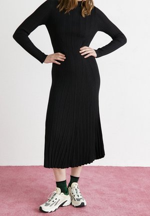 Vrouw in een geribbelde zwarte midi-jurk met lange mouwen, felgroene sokken en beige chunky sneakers, staand op een roze tapijt.