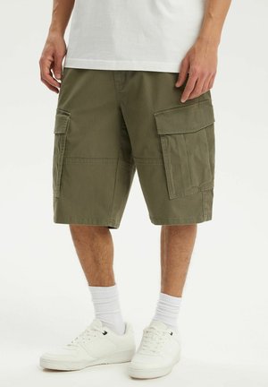 Mężczyzna ubrany w oliwkowe krótkie spodnie typu cargo z dużymi klapkowymi kieszeniami, białe skarpetki, białe trampki i białą koszulkę.