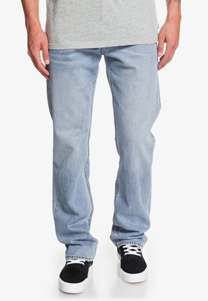 Mann trägt hellblaue Jeans mit geradem Bein, graues T-Shirt und schwarze Sneakers mit weißen Sohlen und Schnürsenkeln, steht mit entspannten Händen.