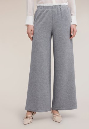 Mujer con pantalones anchos de pierna gris y cintura elástica, y tacones bajos beige de punta puntiaguda, de pie frente a un fondo neutro.