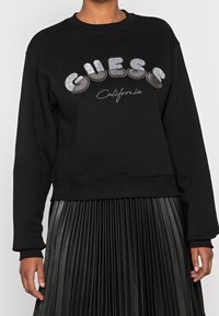 Černá mikina s nápisem "GUESS California" ve stříbrném třpytu. Má klasický kulatý výstřih a zkrácený střih, který je spárován s plisovanou černou sukní.