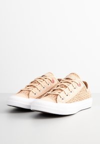 Sneaker in tela beige con una trama intrecciata, suola in gomma bianca, cinque fori per i lacci e un dettaglio rosso sul lato.