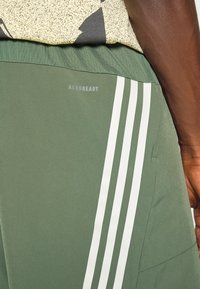 Vihreät urheilushortsit, joissa on valkoiset pystyribbit, valmistettu kevyestä materiaalista ja AEROREADY-logo takana.