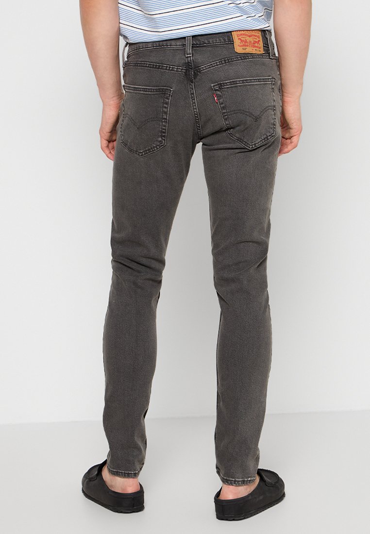 Graue Denim-Jeans mit Slim-Fit, ausgestattet mit Gesäßtaschen und einem Leder-Logo-Patch. Getragen mit schwarzen Slippern.
