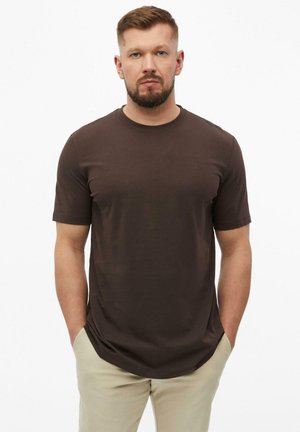 Homme barbu aux cheveux courts portant un t-shirt marron foncé à manches courtes et un pantalon beige clair, debout, les mains dans les poches, sur fond blanc.
