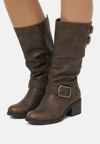 Bottes en cuir marron mi-mollet avec de petits talons carrés, fermeture éclair sur le côté, sangles décoratives autour de la cheville et en haut, portées sur des jambes nues.