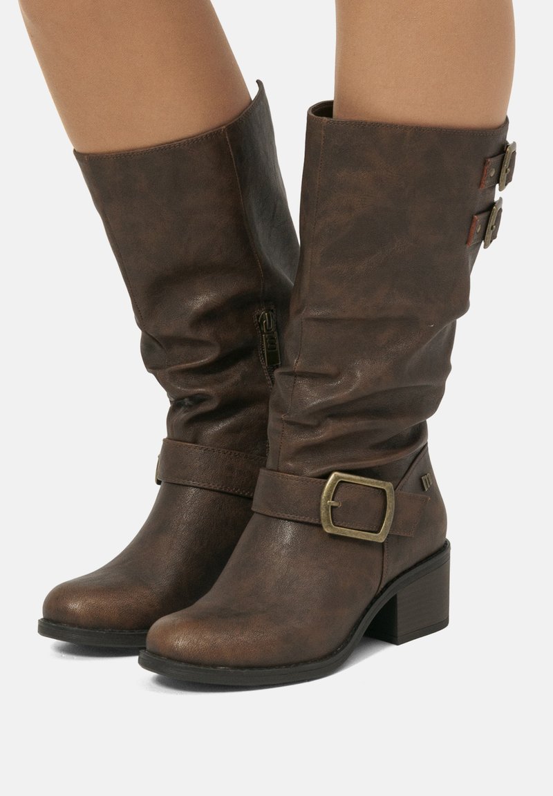 Bottes en cuir marron mi-mollet avec de petits talons carrés, fermeture éclair sur le côté, sangles décoratives autour de la cheville et en haut, portées sur des jambes nues.