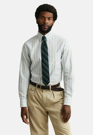 Uomo in piedi che indossa una camicia bianca a righe con bottoni, cravatta a righe blu navy e verde, pantaloni beige plissettati e una cintura intrecciata scura.