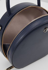 Borsa rotonda in pelle blu navy con una cerniera dorata, caratterizzata da una texture liscia e un manico superiore singolo. Interno foderato con tessuto chiaro.