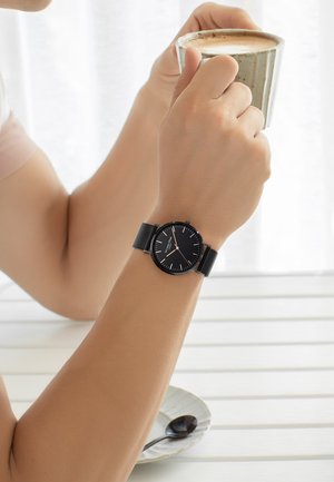 Montre ronde noire avec des index en argent, bracelet en mailles, portée sur un poignet, tenue par une main autour d'une tasse à café beige texturée.