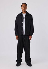 Veste noire à boutons, avec deux poches poitrine, portée sur un t-shirt blanc. Un pantalon noir ample et des baskets noires complètent la tenue.