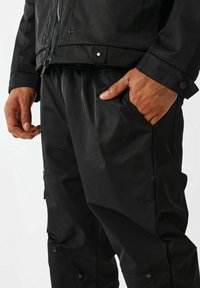Giacca nera con zip e polsini con bottoni, abbinata a pantaloni con tasca laterale, realizzata in un tessuto morbido e impermeabile.
