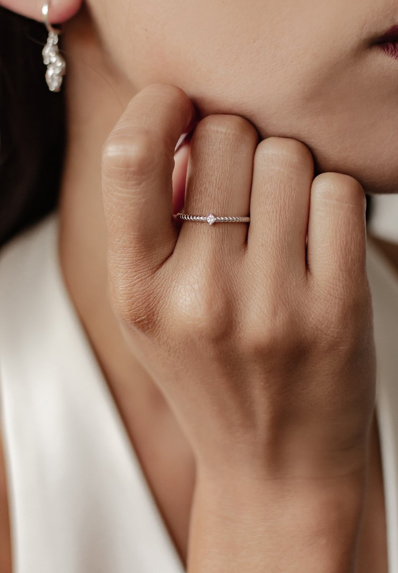 Bague en argent avec un design torsadé, mettant en valeur une petite pierre précieuse ronde. Une main est partiellement visible, reposant sur le menton.