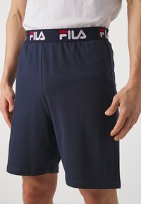 Tumesinine puuvillased lühikesed püksid painduva vöökoha ja valge-punase Fila logo vöökohtades. Sirge lõikega, sile tekstuur.