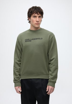 Man draagt een olijfgroen sweatshirt met de tekst "KARL LAGERFELD JEANS", gecombineerd met een zwarte broek, staand tegen een effen witte achtergrond.