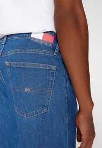 Vaqueros de mezclilla azul con un corte de tiro medio, que incluyen un bolsillo trasero y una etiqueta con bandera en la cintura. La costura es visible a lo largo de los bordes.