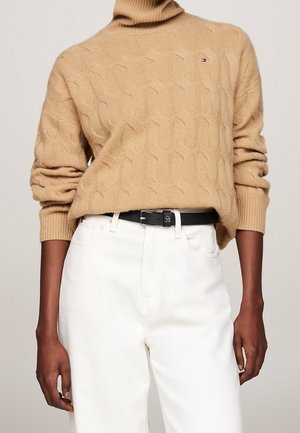 Personne portant un pull à col roulé beige en maille torsadée avec un petit logo et un pantalon blanc taille haute maintenu par une ceinture noire.