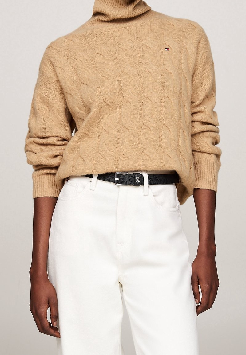 Personne portant un pull à col roulé beige en maille torsadée avec un petit logo et un pantalon blanc taille haute maintenu par une ceinture noire.
