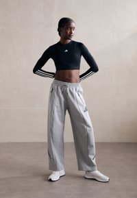 Top corto nero con maniche lunghe e logo Adidas bianco, abbinato a pantaloni della tuta grigi con tasche laterali e dettagli neri. Scarpe da ginnastica bianche.