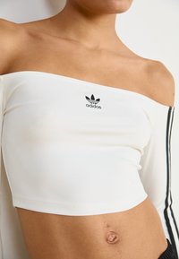 Bijela crop majica s otvorenim ramenima i crnim Adidas logom, s dugim rukavima koji imaju crne vertikalne pruge sa strane. Glatka tekstura tkanine.