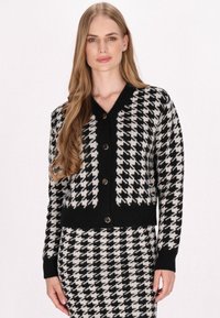 Svetr s houndstooth vzorem v černobílé barvě, s V výstřihem, žebrovanými manžetami a pěti předními knoflíky. Měkký pletený materiál, zkrácený střih.