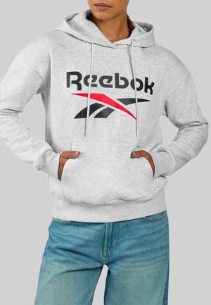 Persoon draagt een lichtgrijze Reebok-hoodie met voorzak en een blauwe spijkerbroek, handen in de zakken gestopt, tegen een effen achtergrond.