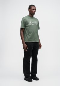 Tricou verde din bumbac cu imprimeu alb, asociat cu pantaloni din denim negri și adidași negri. Design minim, croială standard.