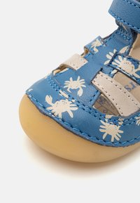 Sininen nahkasandaali, jossa on valkoisia rapukuviota, säädettävä remmi ja vaaleanbeige kumipohja pyöreällä varvaskopalla. Erityinen tikkaus yksityiskohta.