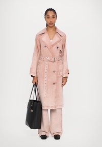 Manteau trench en denim rose avec des boutons double boutonnage, taille ceinturée et détails cloutés ; associé à un sac fourre-tout noir et un pantalon assorti.