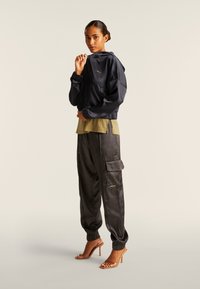 Chaqueta con capucha azul marino y puños elásticos, camisa verde oliva de capas y pantalones cargo negros de satén con bolsillos, combinados con tacones marrones de tiras.